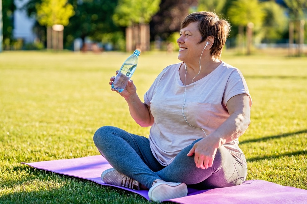 An image of an older woman sitting on a yoga mat drinking a bottle of water suggesting she is embracing the menopause milestone of her life